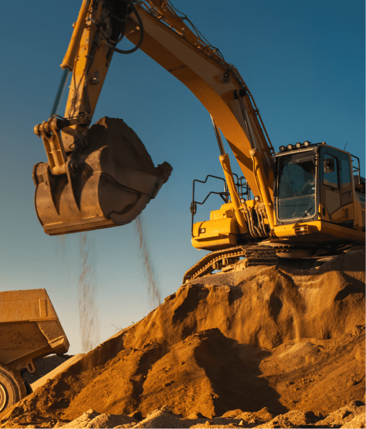 A yellow excavator is positioned on a mound of dirt, using its bucket to transfer soil into a nearby dump truck under a clear blue sky.